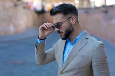 side view of confident elegant man in suit looking down and adjusting sunglasses while posing outside