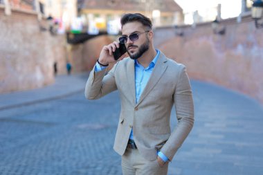 portrait of elegant young businessman in suit having a phone conversation and walking with hand in pockets outside in an old city