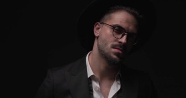close up video of elegant bearded man with hat and eyeglasses looking to side and fixing suit while camera is zooming out with police lights on