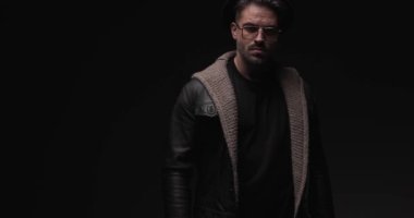 serious young man with glasses wearing wool cardigan and leather jacket, being confident, crossing arms and looking to side