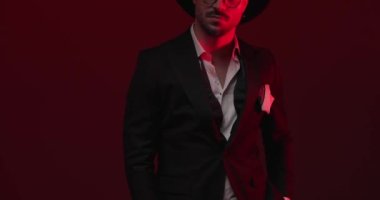 unshaved businessman with hat holding hand in pockets, standing and adjusting eyeglasses, looking to side