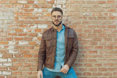 Eski bir ortaçağ kasabasında, gözlüklü ve deri ceketli, elleri vücudunun etrafında serbestçe sallanan yakışıklı sıradan bir adamın portresi.