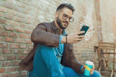 Eski bir ortaçağ kasabasında içki içerken, gözlük ve deri ceket giyerken telefonuyla mesajlaşan yakışıklı bir adamın portresi.