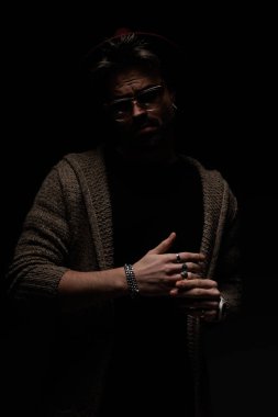 Yüzüklerini düzelten, bordo şapka takan, gözlük takan ve stüdyo arka planında yün ceket giyen sıradan bir gencin portresi.