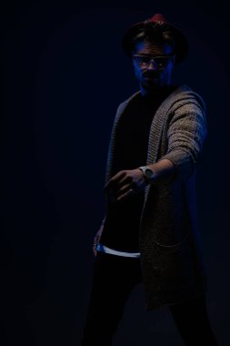 Çekici erkek modelin portresi. Kollarını vücuduna dolamış, koyu renk bir şapka, gözlük ve yün ceket giyiyor.