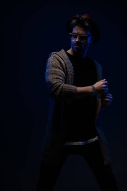 Dramatik ve gizemli bir şekilde poz veren genç moda adamı portresi, koyu renk stüdyo arka planında bordo şapka, gözlük ve yün ceket giyiyor.