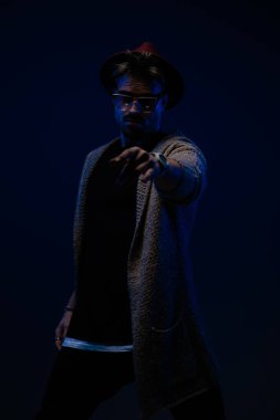 Kamerayı gösteren, koyu renk bir şapka, gözlük ve yün palto takan genç bir modelin portresi.