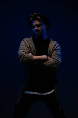 Koyu renk stüdyo arka planında bordo şapka, gözlük ve yün ceket giyen, sert tavırlı yakışıklı erkek modelin portresi.