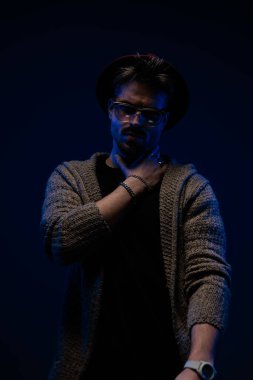 Yakışıklı sıradan bir adamın portresi boynuna piercingli bir şekilde dokunuyor, koyu renk bir şapka, gözlük ve yün ceket giyiyor.