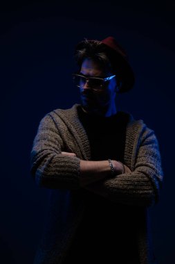 Koyu renk stüdyo arka planında bordo şapka, gözlük ve yün palto giyerek kollarını çapraz geçirip yüzünü çeviren seksi mankenin portresi.