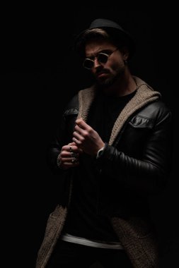 Şapkalı ve gözlüklü seksi adam gözlerini kaçırıyor ve deri ceketi ve yün hırkayı tamir ediyor, havalı ve kendinden emin ve siyah arka planda poz veriyor.