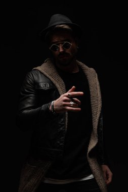 Seksi, gizemli moda adamı elini kaldırıyor ve el hareketi yapıyor, yün hırka, deri ceket, güneş gözlüğü ve şapka giyiyor.