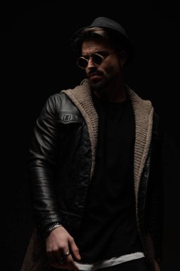 Çekici sakallı genç adam siyah arka planda şapka, güneş gözlüğü, siyah deri ve yün hırka giyiyor.