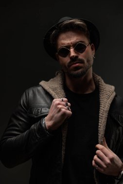 Tıraşsız, güneş gözlüklü, deri ceketli ve yün hırkalı, esrarengiz bir şekilde siyah arka planda poz veren havalı bir adam.