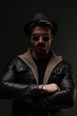Deri ceketli, yün hırkalı, güneş gözlüklü ve siyah şapkalı kendine güvensiz adam kollarını kavuşturuyor.