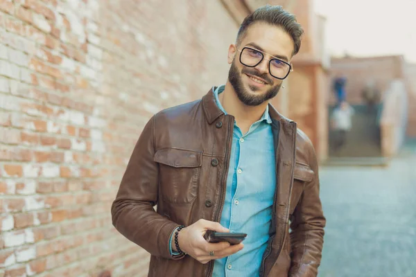若いカジュアルな男の肖像画暖かい笑顔で彼の電話でテキストメッセージ 眼鏡と革のジャケットを身に着けています 古い中世の町の屋外 — ストック写真