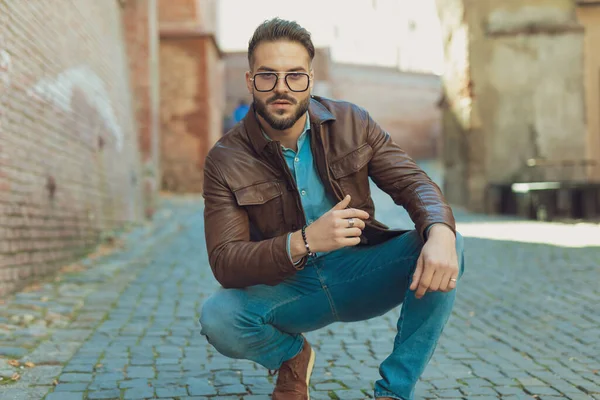 Portrait of young casual man posing with style in a squatted position, wearing eyeglasses and leather jacket, outdoor in an old medieval town