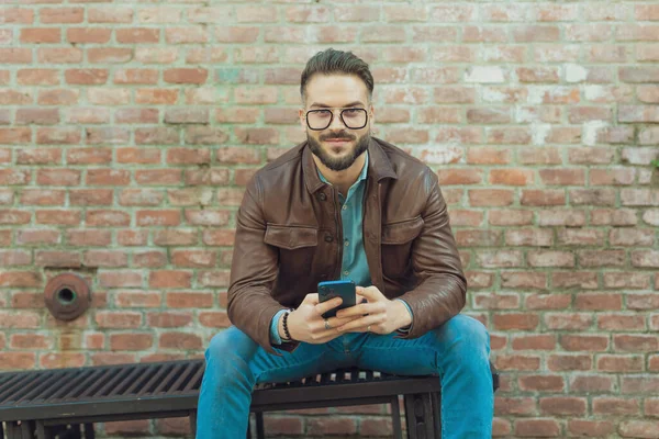 若いカジュアルな男の肖像グリルに座って 彼の電話でテキストメッセージ 眼鏡や革のジャケットを着て 古い中世の町の屋外 — ストック写真