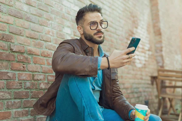 在一个中世纪的古城 一个迷人的休闲男子穿着眼镜和皮夹克 在户外用手机发短信 品尝苏打水的画像 — 图库照片