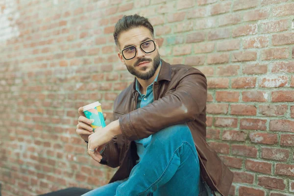 コーヒーを飲みながらベンチでリラックスする若いカジュアルな男の肖像画 眼鏡と革のジャケットを身に着けて 古い中世の町の屋外 — ストック写真