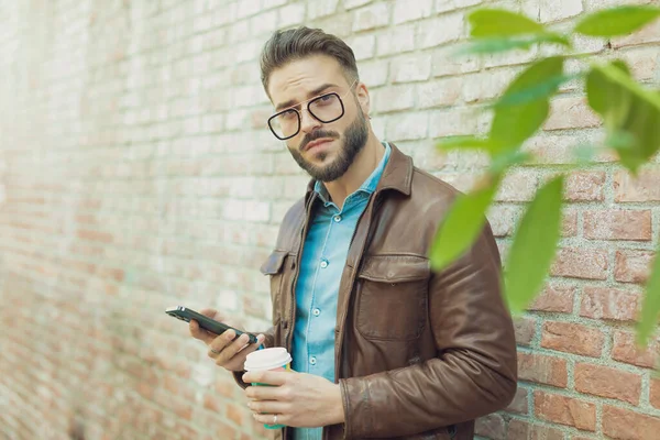 在一座中世纪的古城里 一个穿着眼镜和皮夹克 在户外用电话发短信 靠着墙喝酒的有魅力的休闲男子的画像 — 图库照片