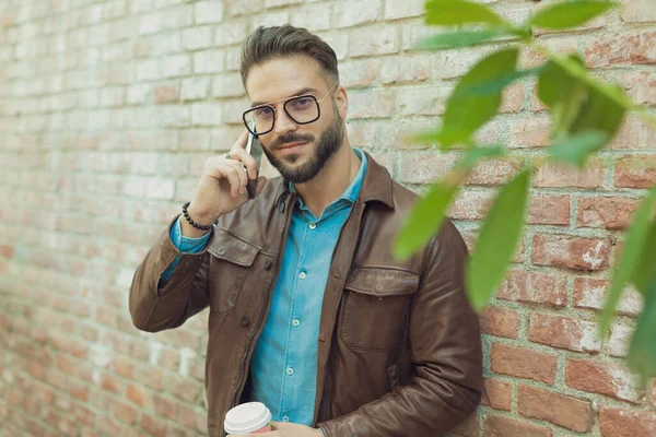 壁に酒を飲みながら電話で話すセクシーなカジュアルな男の肖像画 眼鏡と革のジャケットを身に着けて 古い中世の町の屋外 — ストック写真
