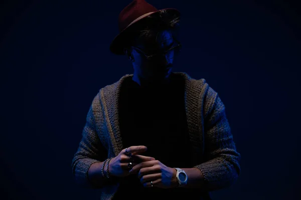 stock image Portrait of attractive fashion man with mysterious vibe posing in dramatic stance, wearing a burgundy hat, eyeglasses and wool coat in dark studio background