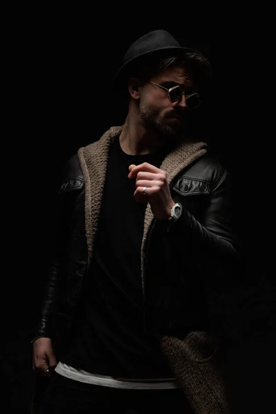 cool fashion man with hat and sunglasses wearing black leather jacket and wool cardigan, looking to side and posing in a cool way on black background
