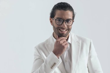 Beyaz takım elbiseli yakışıklı bir adamın portresi. Çeneye dokunuyor, düşünüyor ve gri arka planda gülümsüyor.