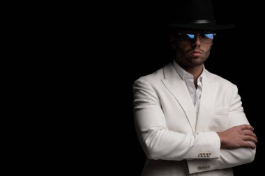 Şapkalı ve yakalı şık bir adam kollarını kavuşturup siyah arka planda poz veriyor.