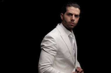 Beyaz ceketi ayarlayan ve siyah arkaplan önünde poz veren zarif genç adamın yan görüntüsü