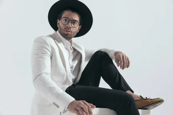 时尚男子 头戴帽子 戴着眼镜 坐在灰色背景前 膝盖上有肘部 — 图库照片
