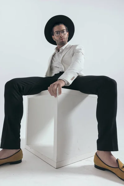 一个头戴帽子 戴着眼镜 很酷的年轻人 坐在白色的木制桌子上 自信地在灰色的背景上摆姿势 — 图库照片