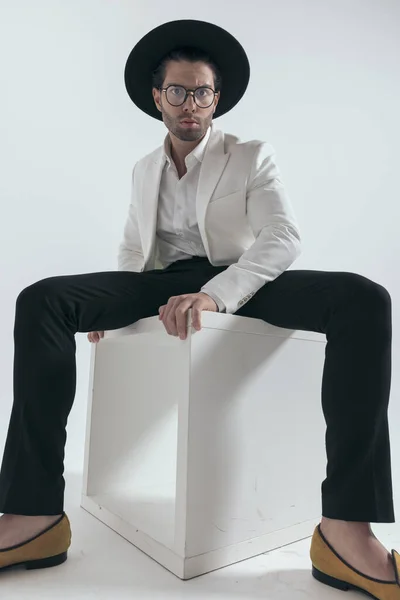 sexy fashion man wearing hat and glasses and posing in a cool way while sitting on white background
