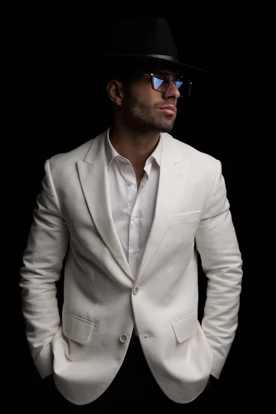 身穿白色夹克套装 头戴帽子 戴着眼镜 开领衬衫的性感时尚男人 双手插在黑色背景的口袋里 — 图库照片
