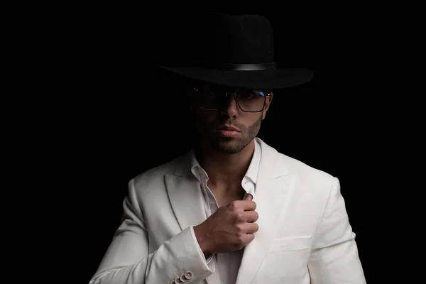 attractive young man wearing hat and glasses and adjusting open collar shirt in front of black background