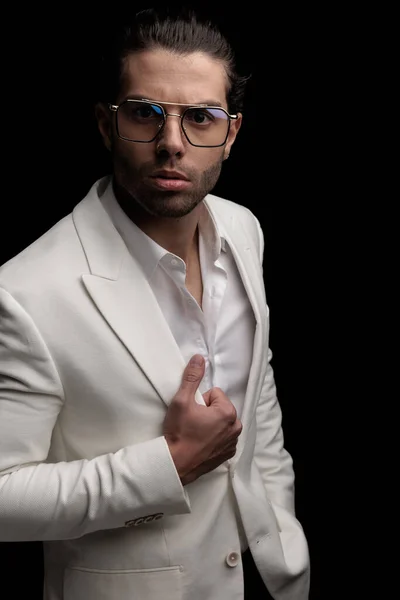 attractive elegant man with open collar shirt adjusting white suit jacket and posing in a cool way in front of black background