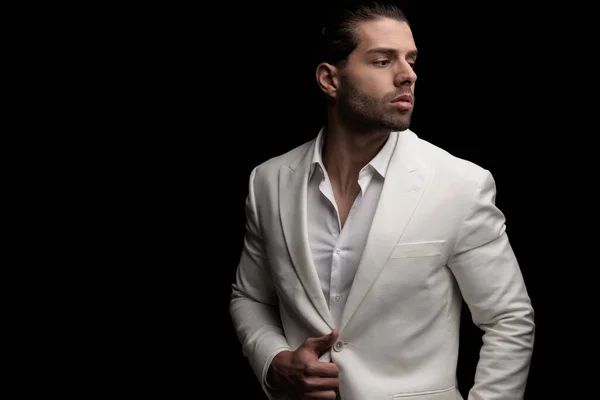 cool elegant man with open collar shirt adjusting and unbuttoning suit while looking to side in front of black background