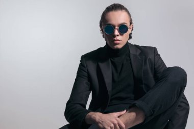 Yakışıklı iş adamının bir bacağını üst üste koyup gri stüdyo geçmişine karşı güzel bir kıyafet giydiği bir moda fotoğrafı.