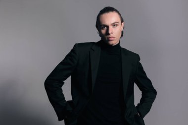 Çekici iş adamının cepleri ve dikkatsizliğiyle poz verdiği ve gri stüdyo geçmişine karşı güzel bir kıyafet giydiği bir moda fotoğrafı.