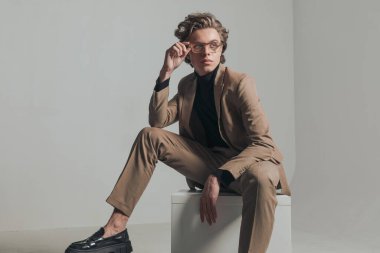 Gri arkaplan önünde diz çökmüş otururken, gözlük takan sarı saçlı moda genç adam.