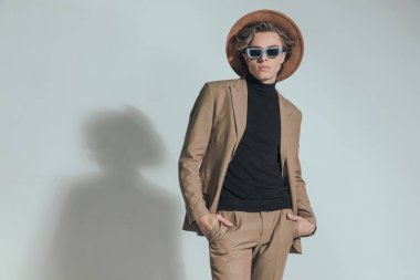 Stüdyoda gri arka planda el ele tutuşan şapkalı ve güneş gözlüklü seksi moda adamı.