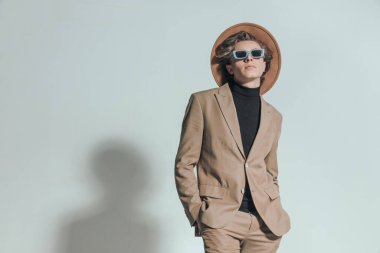 Şapkalı, güneş gözlüklü, el ele tutuşup gri arka planda poz veren havalı bir genç.