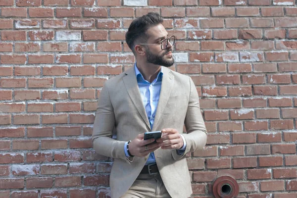 Portrait of young businessman texting on phone while leaning on brick wall and wearing eyeglasses outdoor, in an old medieval town
