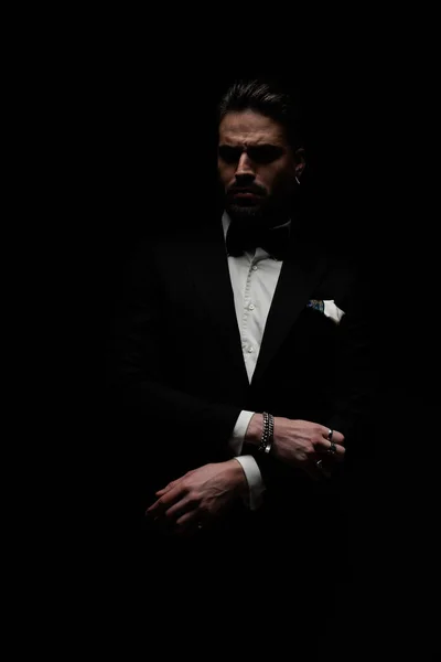 bearded young man in elegant tux holding arms in fashion pose and looking away in front of black background in studio