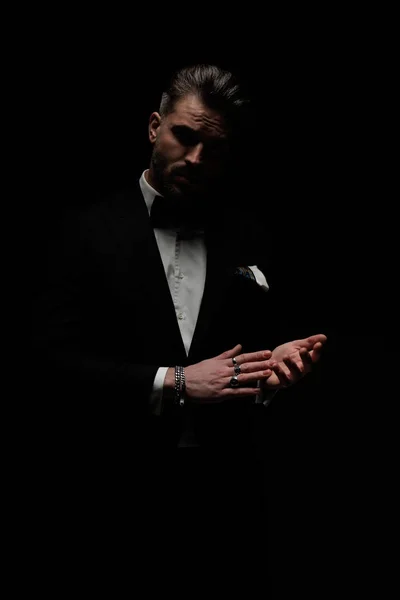 elegant young groom wearing black tuxedo with bowtie and handkerchief rubbing palms and posing in front of black background