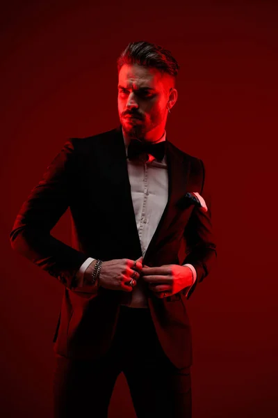 Элегантный Человек Бородой Глядя Сторону Застегивая Черный Смокинг Позируя Красными — стоковое фото