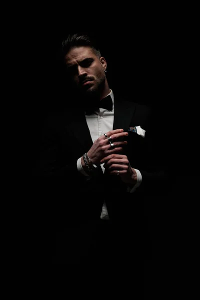 stylish unshaved groom in black elegant tuxedo touching fingers and posing in front of black background in studio