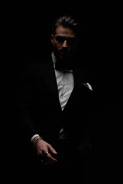 bearded young man in black tuxedo posing in a mysterious way in front of black background