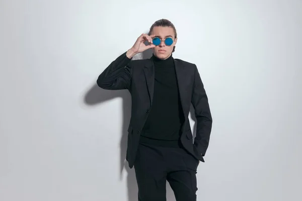 Fashion picture of attractive businessman fixing sunglasses with a hand in pocket and wearing a nice outfit against gray studio background